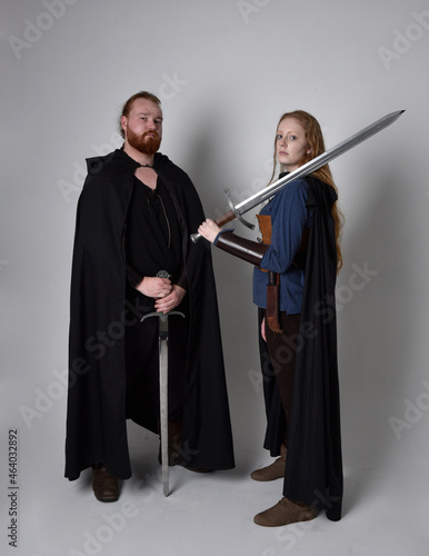 Full length portrait of red haired couple, man and woman wearing medieval viking inspired fantasy costumes, standing fighting pose holding sword weapons, isolated on white studio background.