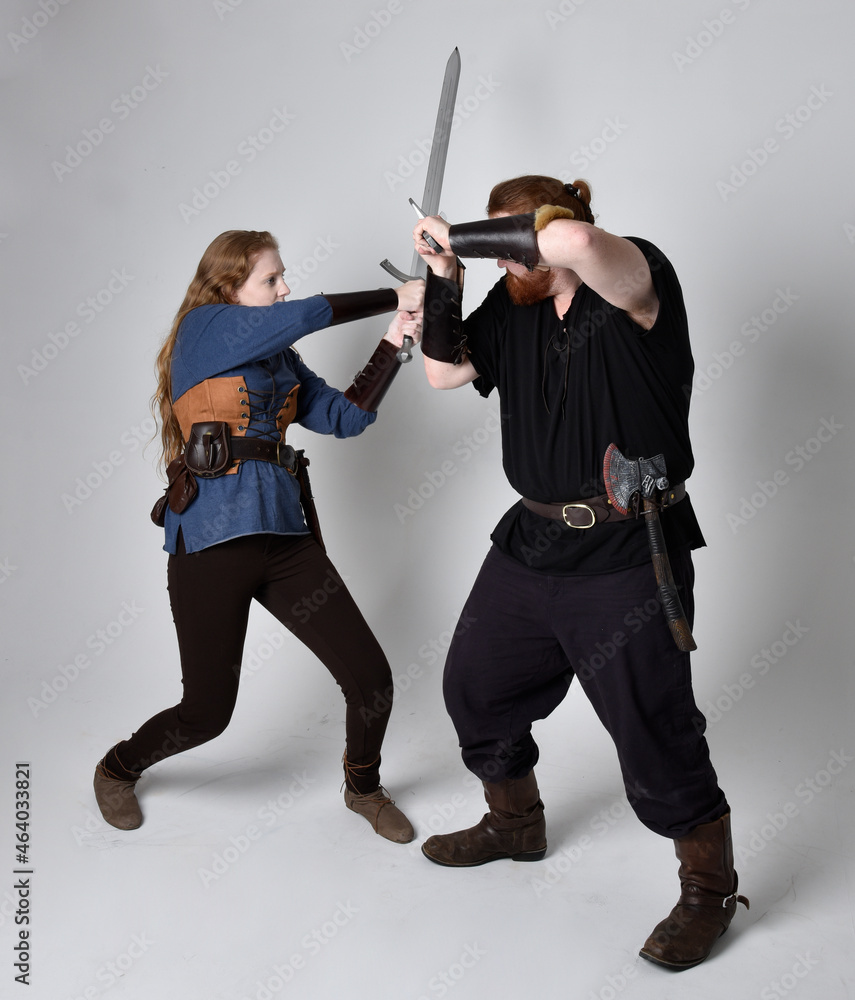 Full length portrait of red haired couple, man and woman wearing medieval viking inspired fantasy costumes, standing fighting pose holding sword weapons, isolated on white studio background. Full length portrait of red haired couple, man and woman wearing medieval viking inspired fantasy costumes, standing fighting pose holding sword weapons, isolated on white studio background.