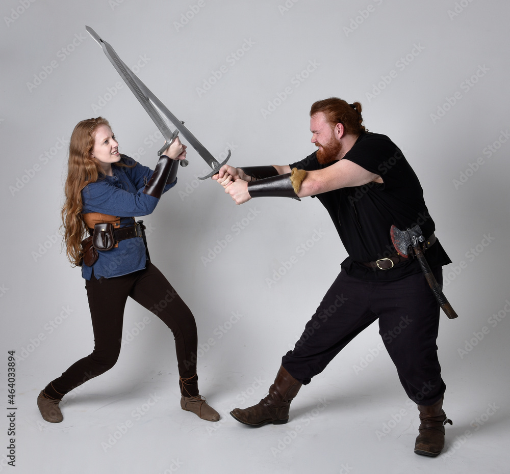 Full length portrait of red haired couple, man and woman wearing medieval viking inspired fantasy costumes, standing fighting pose holding sword weapons, isolated on white studio background. Full length portrait of red haired couple, man and woman wearing medieval viking inspired fantasy costumes, standing fighting pose holding sword weapons, isolated on white studio background.
