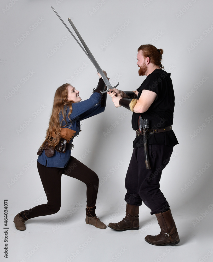 Full length portrait of red haired couple, man and woman wearing medieval viking inspired fantasy costumes, standing fighting pose holding sword weapons, isolated on white studio background. Full length portrait of red haired couple, man and woman wearing medieval viking inspired fantasy costumes, standing fighting pose holding sword weapons, isolated on white studio background.