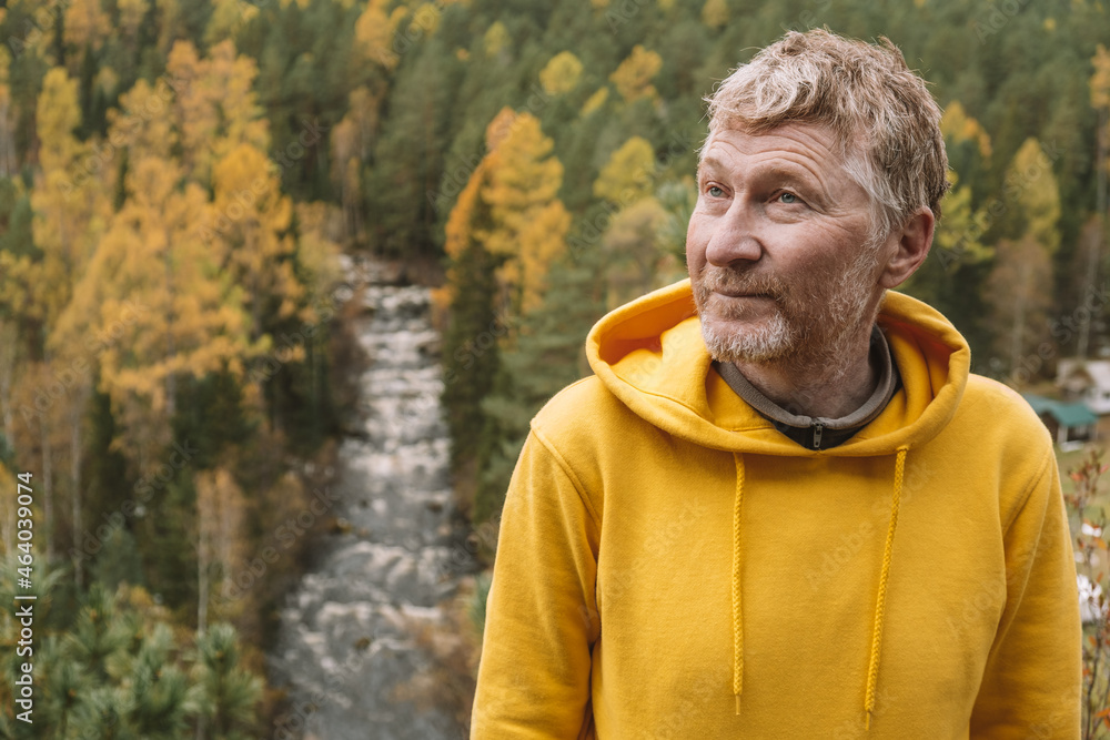 An authentic portrait of a handsome unshaven adult male traveler in a yellow hoodie on top of a mountain, against the background of an autumn forest. Selective focus. Close-up. Sustainable environment An authentic portrait of a handsome unshaven adult male traveler in a yellow hoodie on top of a mountain, against the background of an autumn forest. Selective focus. Close-up. Sustainable environment