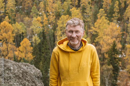 An authentic portrait of a smiling unshaven male traveler in a yellow hoodie on top of a mountain, against the background of an autumn forest. Selective focus. Close-up. Sustainable environment.