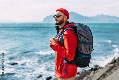 A bearded traveler with a backpack on the background of the sea. Portrait of a traveler in red clothes and sunglasses. A tourist with a backpack stands against the background of the sea. Copy space