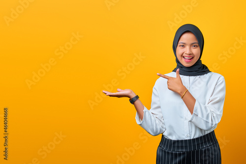 Cheerful young Asian woman presenting product and pointing with fingers on yellow background
