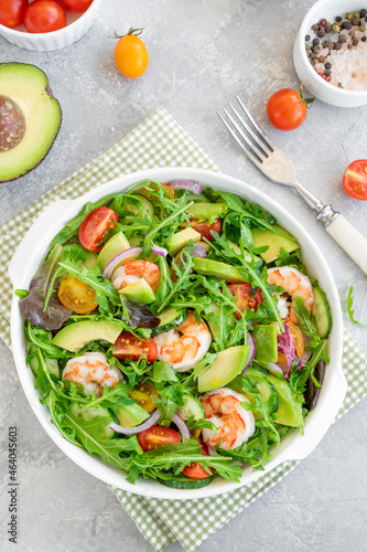 Delicious fresh salad with prawns, tomatoes, avocado, cucumber and onions in a bowl on a gray concrete background. Healthy dish. Top view, copy space.