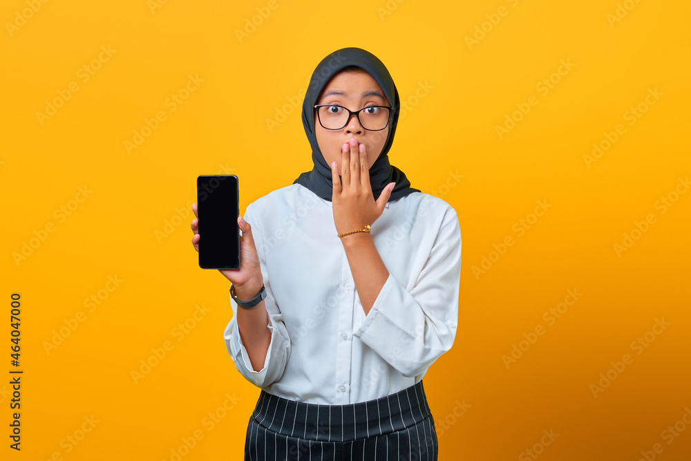 Surprised young Asian woman showing mobile phone blank screen isolated over yellow background Surprised young Asian woman showing mobile phone blank screen isolated over yellow background