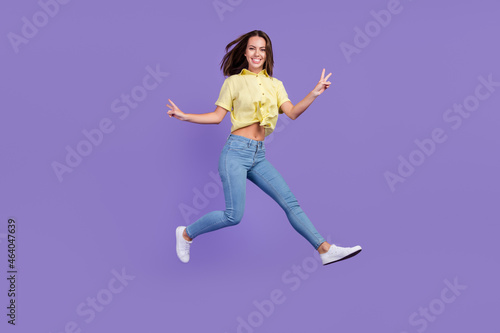 Full length photo of cool millennial brunette lady run show v-sign wear yellow top jeans footwear isolated on violet color background