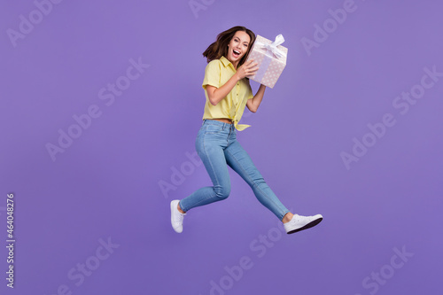 Full length photo of impressed millennial brunette lady jump hold present wear yellow top jeans sneakers isolated on violet color background