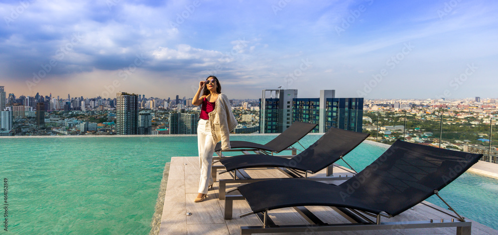 Luxury Asian CEO woman entrepreneur standing on the rooftop swimming pool looking to the sun with skyscraper and cityscape on the background with copy space