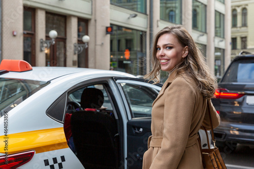 A beautiful young woman gets into a yellow taxi in the city. Smiling blonde in a beige coat. Travel and traffic in the metropolis.