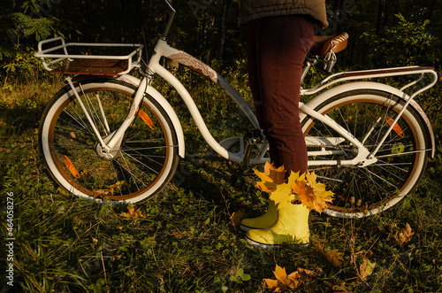 Bicycle with basket in autumn forest with woman standing in yellow rubber boots, filled with yellow leaves.