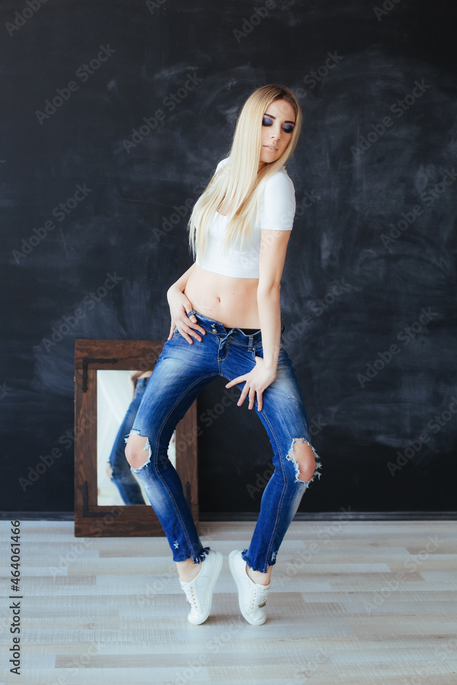 Young blonde woman with vivid makeup in white top and torn blue jeans posing near the mirror