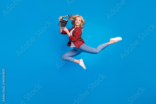 Photo of adorable funky young lady wear plaid shirt smiling humping high holding clapperboard isolated blue color background