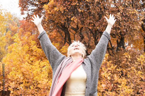 Happy senior woman with hands up standing in autumn park. Adult woman smiling looks up with raised hands. Yellow trees on background. Retirement, elderly health, life insurance, free breathing concept