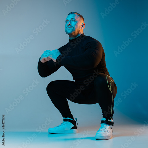 Stylish brutal man doing squat exercises. Athlete workout trending neon blue background. Black sportswear. Fitness gym. Aerobic exercise. Strong muscular guy with textural muscles