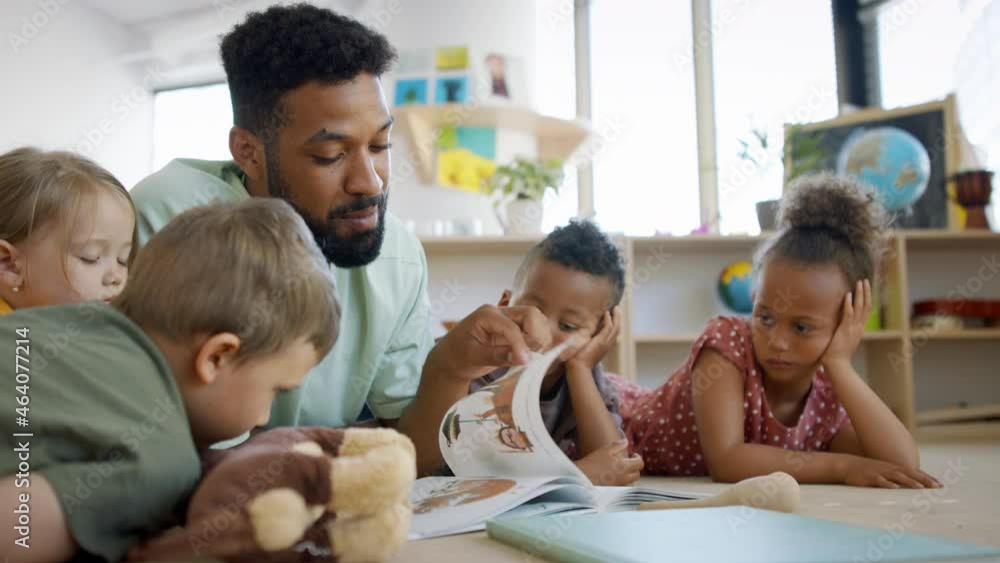 Group of small nursery school children with man teacher on floor indoors in classroom, reading book.