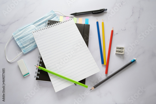 Blank spiral notepad mock up with copy space, crayons, surgical face mask against covid-19 infection and stationary supplies on a marble surface, high angle view from above