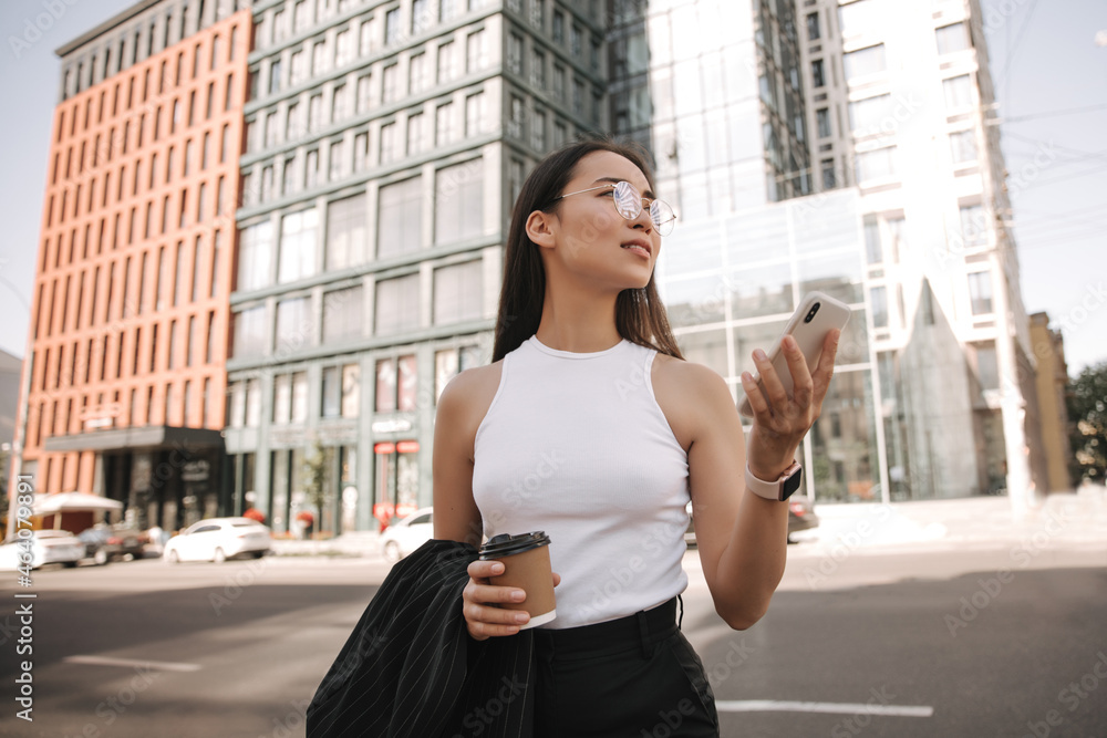 Calm young woman using double gis on her phone looking for right street. She is wearing black suit, round glasses and fragrant coffee in her hand. Telephone communication technology concept