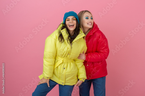 two attractive girl friends active women posing on pink background in colorful winter down jacket of bright red and yellow color having fun together, warm coat fashion trend, sincerely laughing