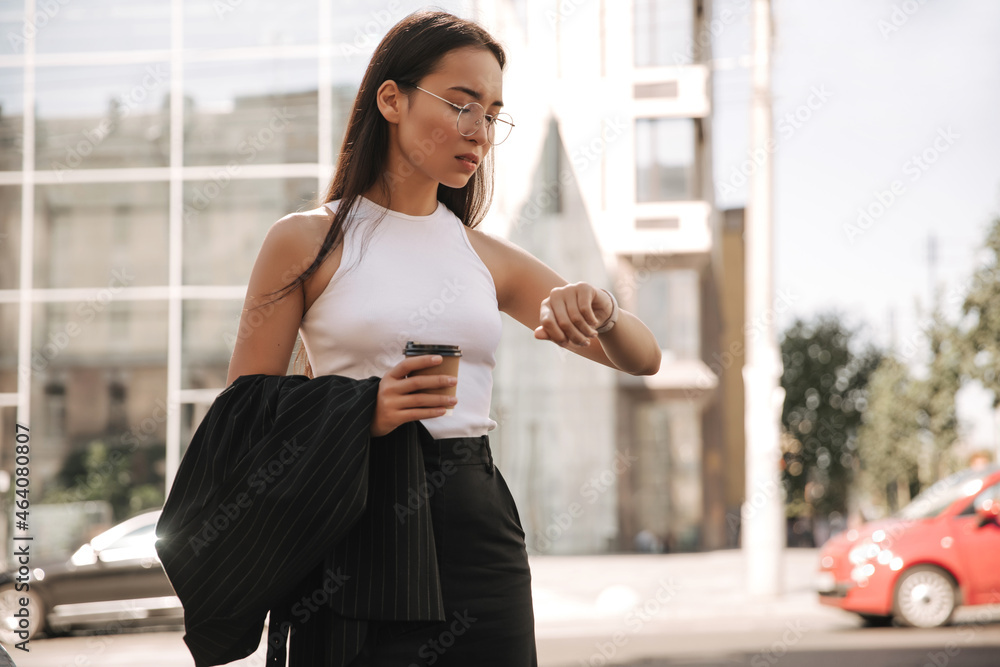 Not full-length beautiful young asian brunette looks upset at time near panoramic house. Woman in glasses with coffee is looking forward to meeting with colleagues at work. 