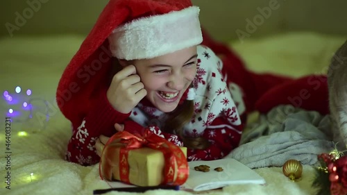 A teenage girl in Santa's red hat and red christmas jacket lies on the bed and writes a letter to Santa Claus. Near the British cat. New Year. hildren's Christmas. Atmosphere. Home. photo