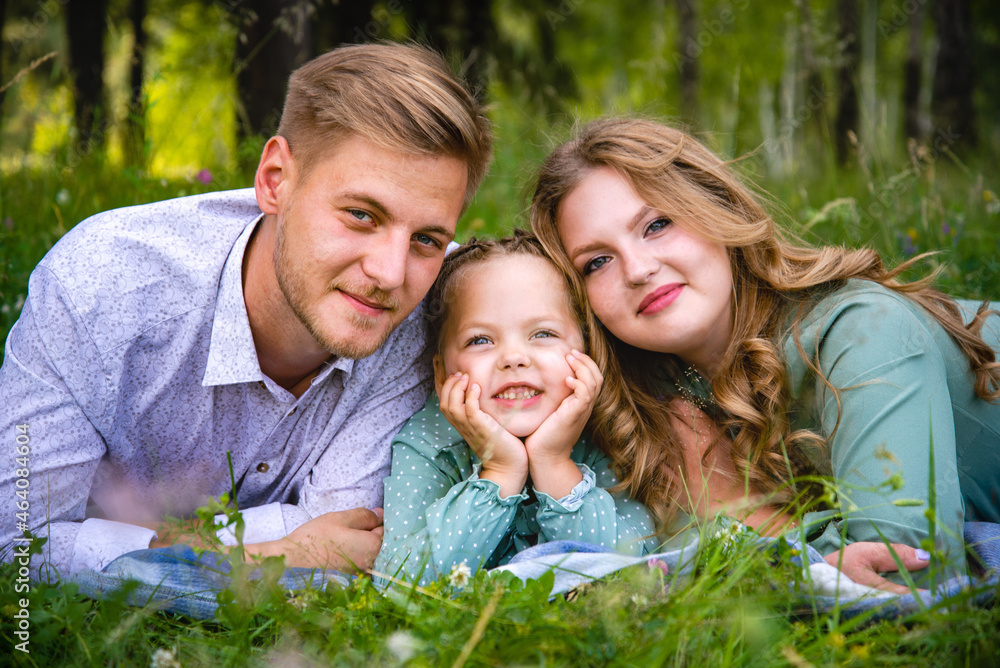 Fototapeta premium Happy young family father, mother and little daughter smiling as they lie on the green grass on a sunny day