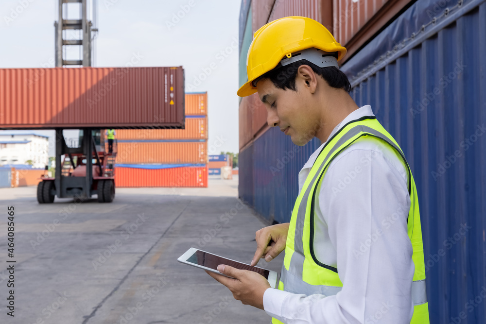 Engineer or foreman holding tablet and looking down to checking inventory or job details with cargo container background. Engineering site and working with technology concept. Engineer or foreman holding tablet and looking down to checking inventory or job details with cargo container background. Engineering site and working with technology concept.