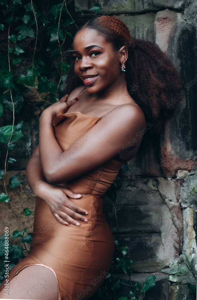 Fototapeta premium Beautiful African black woman walking in park in autumn. Beautiful young woman with brown skin. Smiling black woman with afro hair wearing brown dress with high slit.