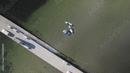 Aerial view of surfers sitting on their surfboards in the water next to a pier on a sunny day. 4K drone video.