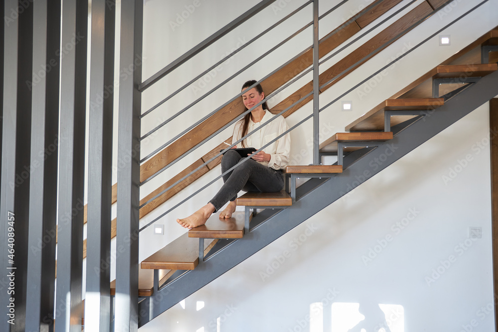 Woman working from home in her living room while sitting on staircase. Modern metal stairway in interior design Woman working from home in her living room while sitting on staircase. Modern metal stairway in interior design