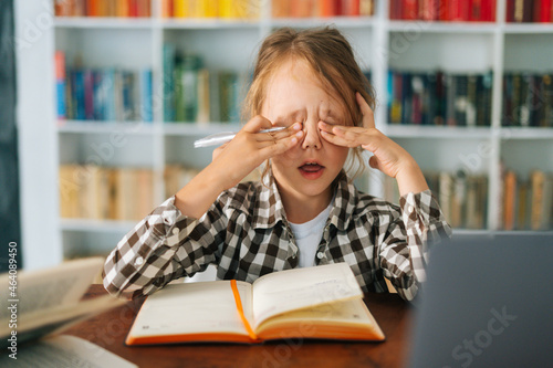 Quadro em tela Close-up front view of frustrated exhausted primary child school girl rubbing tired from active reading eyes, doing lots homework sitting at desk with workbook, selective focus