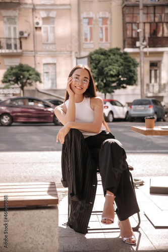 Young asian woman enjoying coffee sitting on terrace of cafe on street of old city in sunny day. She is wearing stylish black suit, sandals with heels. Urban life concept © Look!