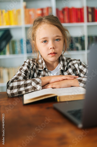 Vertical portrait of focused primary child school girl sitting at table with laptop and paper book in cozy children room, looking at camera. Cute pupil schoolgirl e-learning online using computer.
