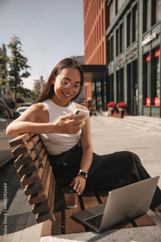 Beautiful young asian woman resting on bench browses social networks in phone. Works at laptop, drinks coffee. sophisticated lady is dressed in white t-shirt, black trousers.