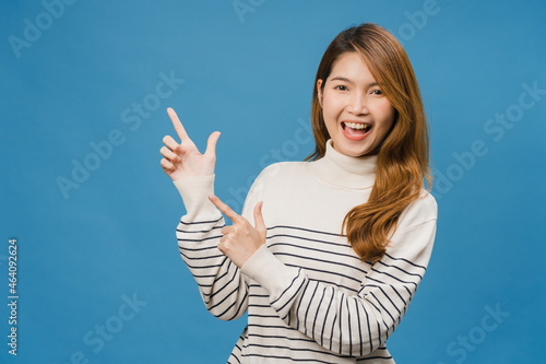 Portrait of young Asian lady smiling with cheerful expression, shows something amazing at blank space in casual clothing and looking at camera isolated over blue background. Facial expression concept.