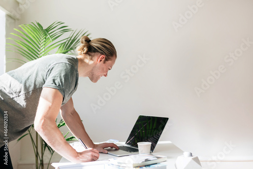 Blond young designer leaning over a desk looking at a laptop screen photo