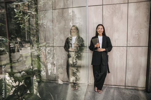 Fototapeta Naklejka Na Ścianę i Meble -  Attractive young asian brunette with her hair down in full length is standing indoors. She is wearing black business suit, folder with documents in her hands. Confidence in their work, concept.