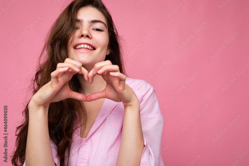 Photo of young positive happy smiling beautiful wavy-haired brunette woman with sincere emotions wearing trendy pink shirt isolated over pink background with free space and showing heart shape with Photo of young positive happy smiling beautiful wavy-haired brunette woman with sincere emotions wearing trendy pink shirt isolated over pink background with free space and showing heart shape with