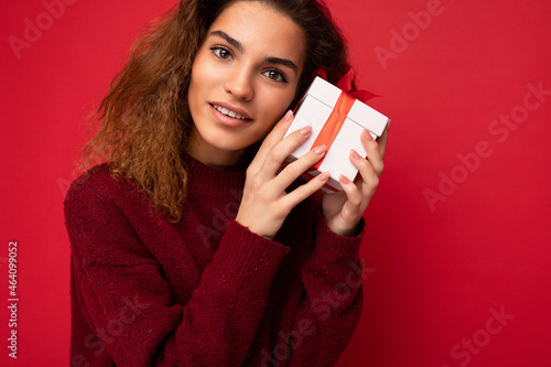Closeup photo of attractive happy joyful young brunette curly female person standing isolated on red background wall wearing red sweater holding gift box with red ribbon looking at camera