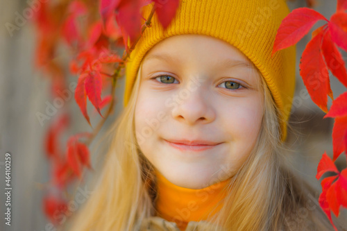 portrait of a smiling beautiful little blonde girl with long flowing hair in a yellow hat against a background of colorful vine leaves