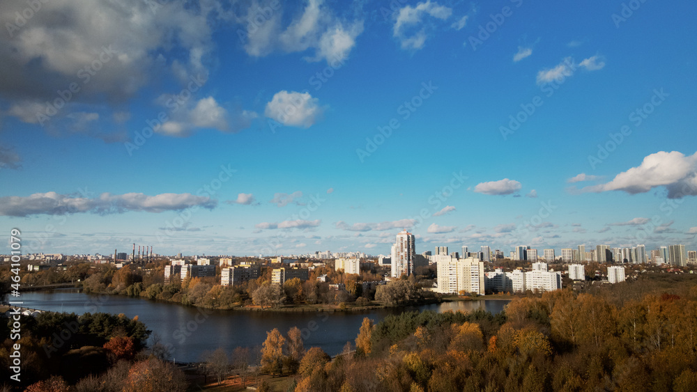 Flight over the autumn park. Park on the shore of a large lake. On the horizon there is a blue sky and city houses. Trees with yellow autumn leaves are visible.  Aerial photography.