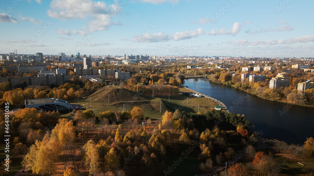Flight over the autumn park. Park on the shore of a large lake. On the horizon there is a blue sky and city houses. Trees with yellow autumn leaves are visible.  Aerial photography.