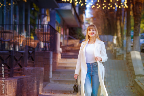 Portrait of a beautiful business woman walking outdoor. Girl weared in a white woman coat, jeans. City street autumn.