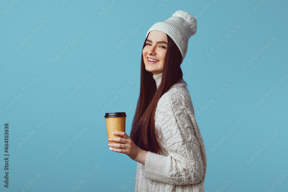 Happy young woman wearing white oversize sweater and hat isolated over blue background, holding takeaway coffee Happy young woman wearing white oversize sweater and hat isolated over blue background, holding takeaway coffee
