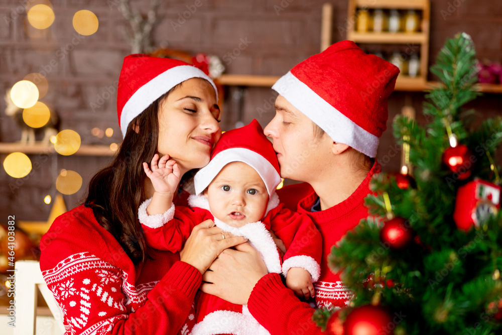 New Year or Christmas, a happy young family mom, dad and baby in the dark kitchen of the house with a Christmas tree in red sweaters and Santa Claus hats smiling, hugging congratulating on the holiday New Year or Christmas, a happy young family mom, dad and baby in the dark kitchen of the house with a Christmas tree in red sweaters and Santa Claus hats smiling, hugging congratulating on the holiday