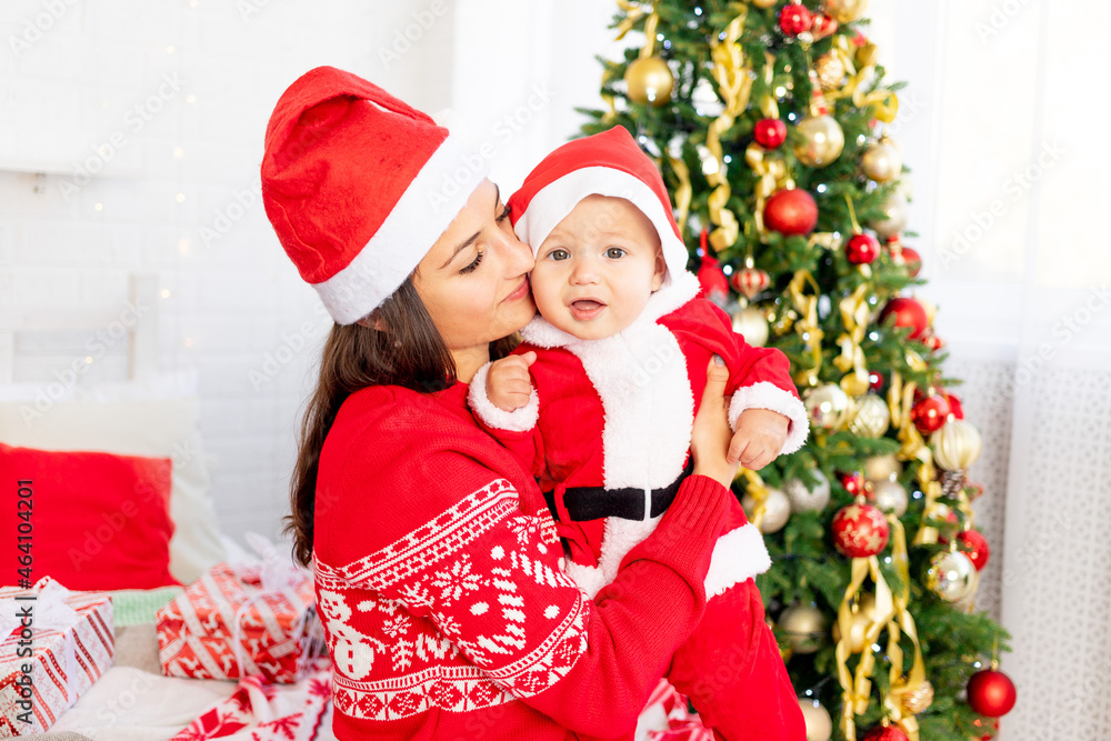 New Year or Christmas, a young mother with a baby on the bed at home by the Christmas tree in a Santa Claus costume hugging and waiting for the holiday smiling New Year or Christmas, a young mother with a baby on the bed at home by the Christmas tree in a Santa Claus costume hugging and waiting for the holiday smiling