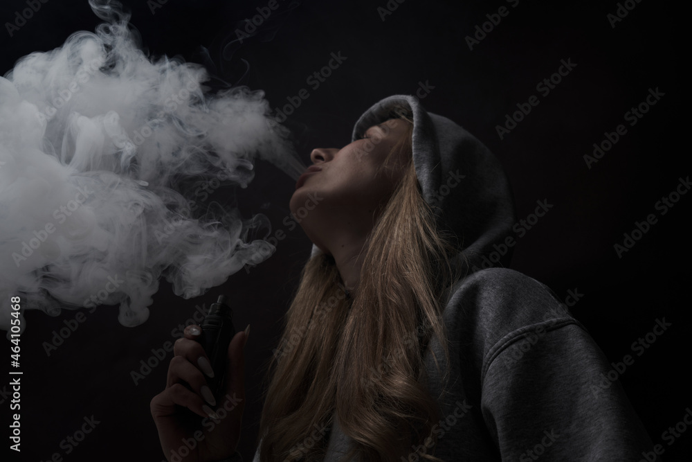 Portrait of beautiful blonde girl in gray hoodie smokes vape isolated on black studio background, cloud of steam smoke, mini hookah