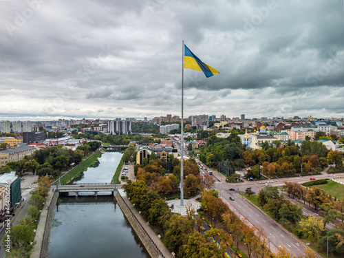 Tallest flag of Ukraine, flagpole with epic gray cloudscape, city aerial view above river Lopan embankment near Svyato-Pokrovskyy Monastyr in Kharkiv, Ukraine