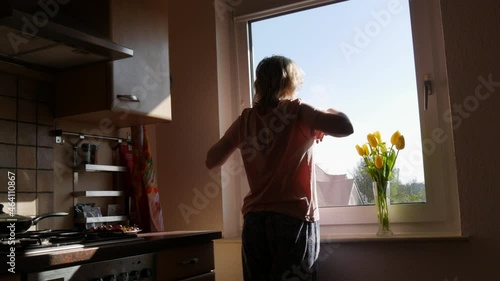 Young woman just woke up, stretching herself sleepy in pajamas, comes to window and looks into it, on the windowsill there is a bouquet of yellow tulips in a vase, inhales the aroma. Dutch corner photo