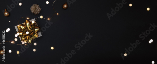 Christmas composition. White gift box with golden ribbon, New Year balls and sparkling lights garland in xmas composition on black background for greeting card. Flat lay, top view, copy space.